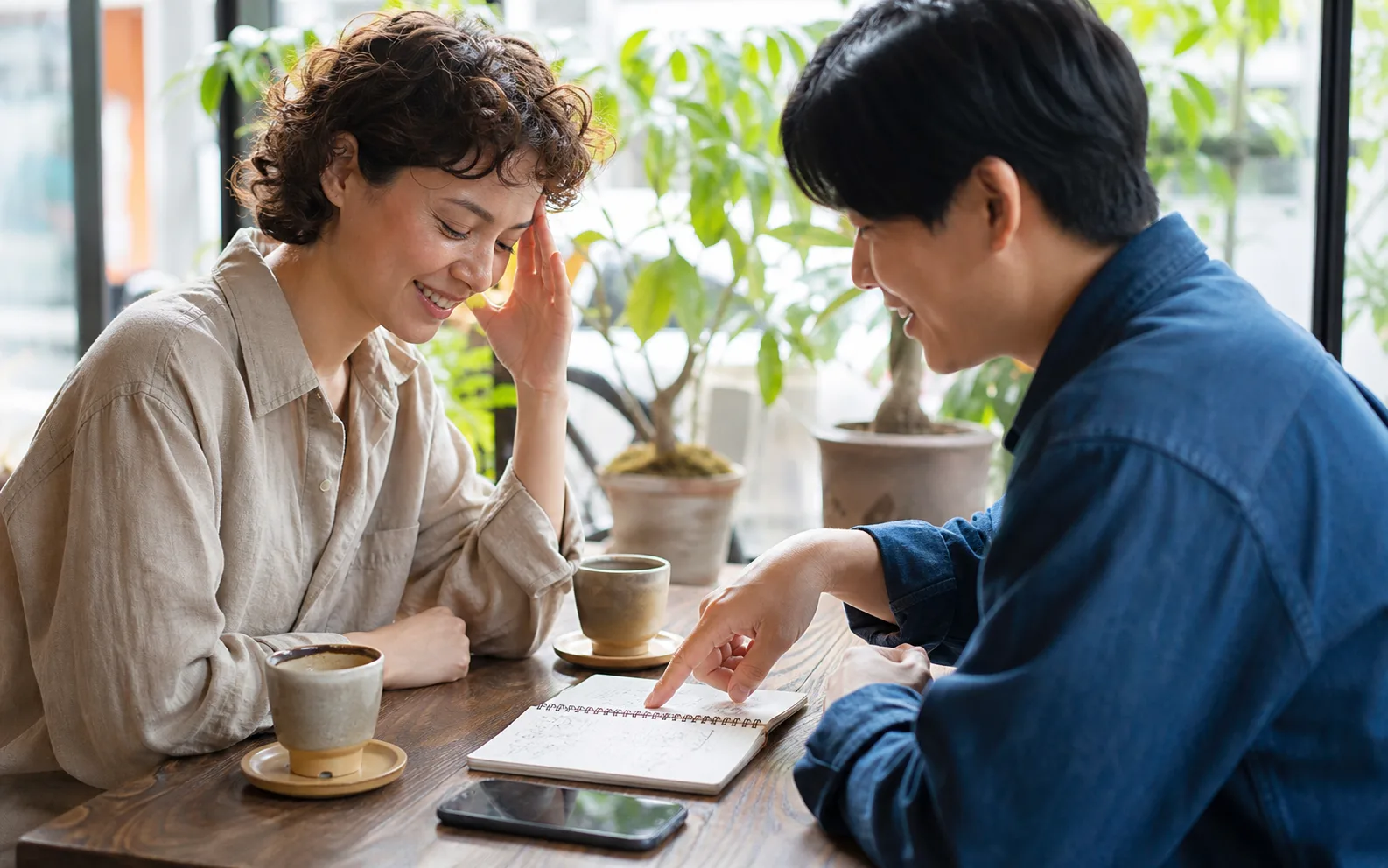 Two people smiling during a cafe conversation while looking at a notebook and phone together.
