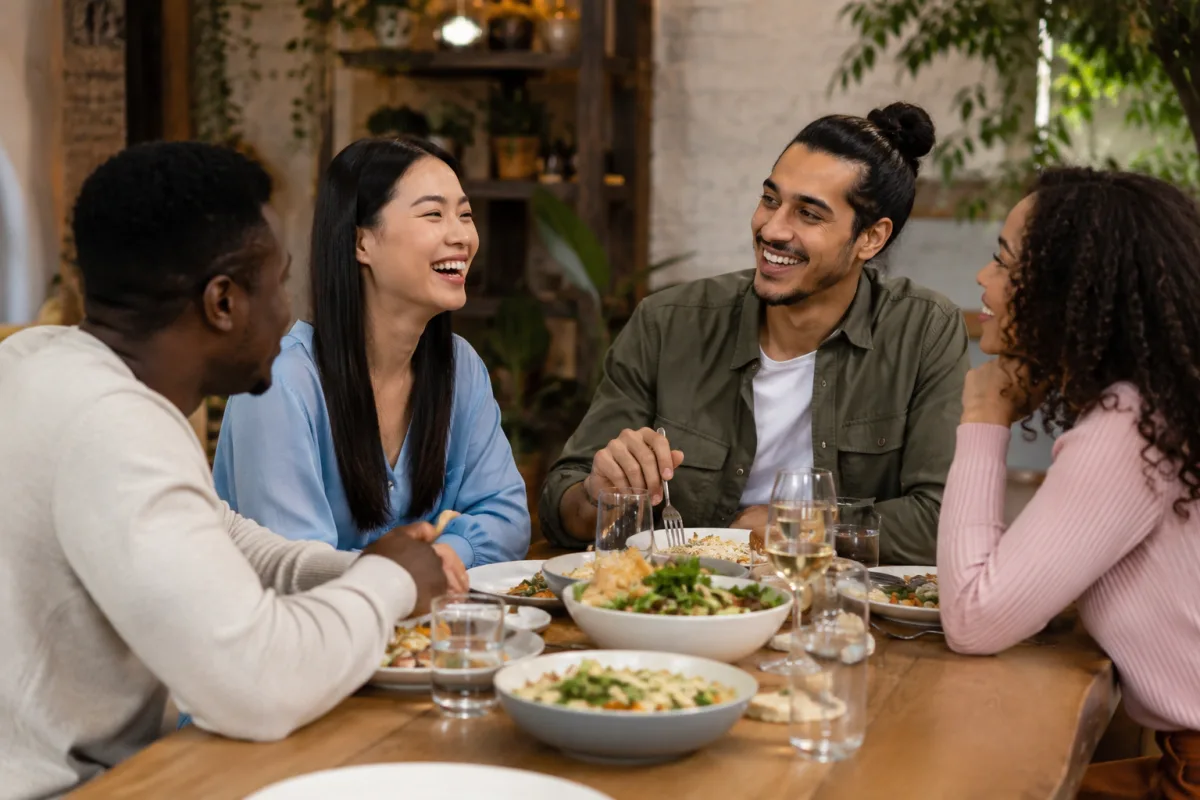 A realistic photo of four friends from different racial backgrounds sitting around a dining table in a cozy restaurant setting, smiling and talking together while sharing food. The group includes Black, East Asian, South Asian, and mixed-race individuals, creating a warm and multicultural atmosphere with soft lighting and a relaxed social vibe.