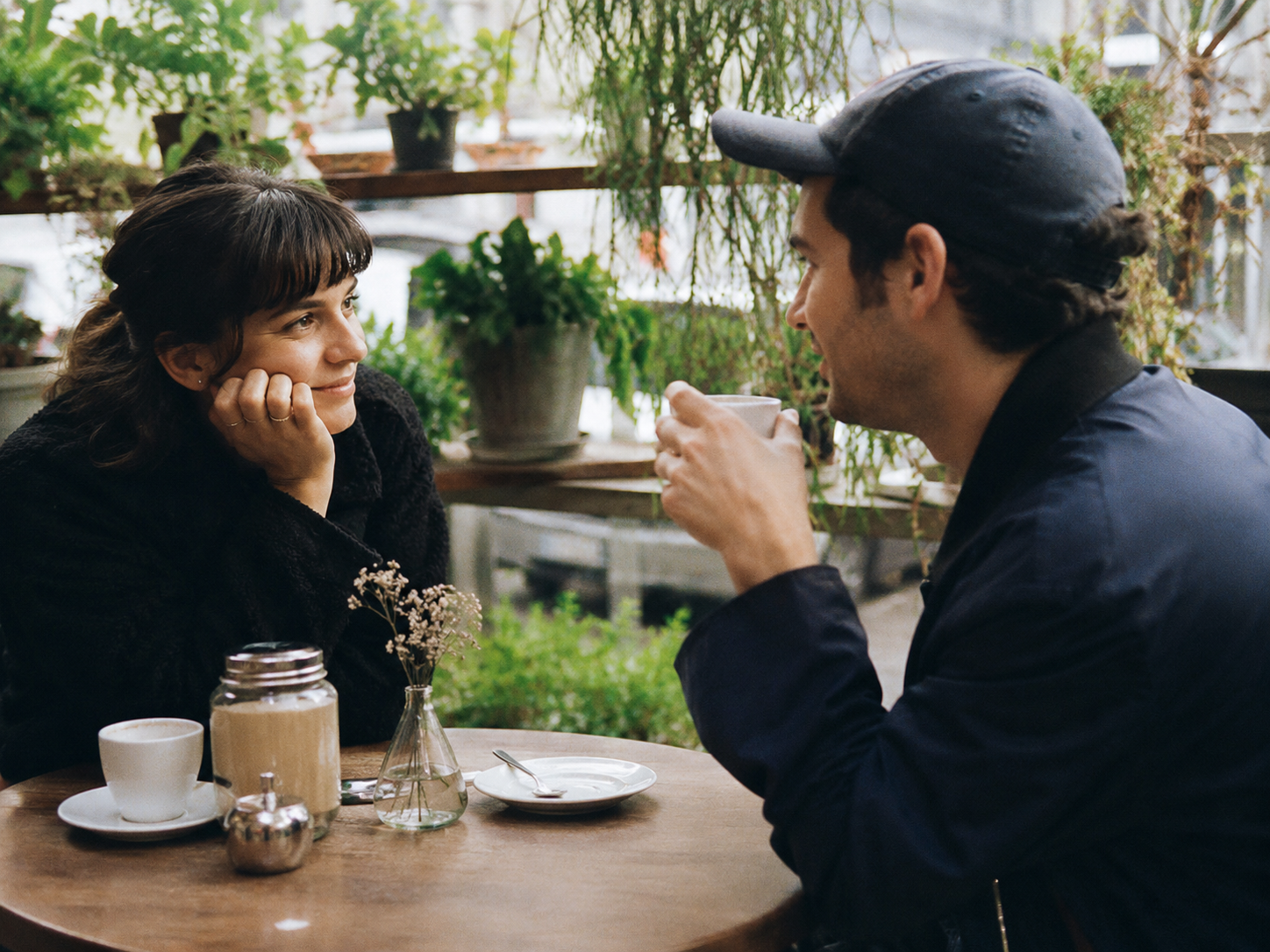 Two people having a relaxed conversation at a cafe table, surrounded by plants and greenery, with coffee cups on the table in a candid analog-style photo.